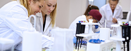 På billedet ses to elever iført hvide kitler, der foretager et eksperiment i et lokale med andre elever.