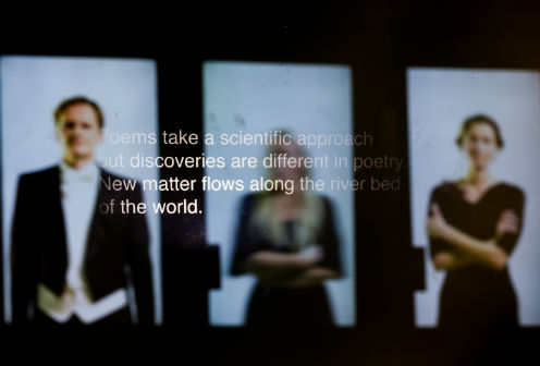 The caption is taken from the event. A blurry picture of three people in the background and in front the following text: "Poems take a scientific approach but discoveries are different in poetry. New matter flows along the river bed of the world."